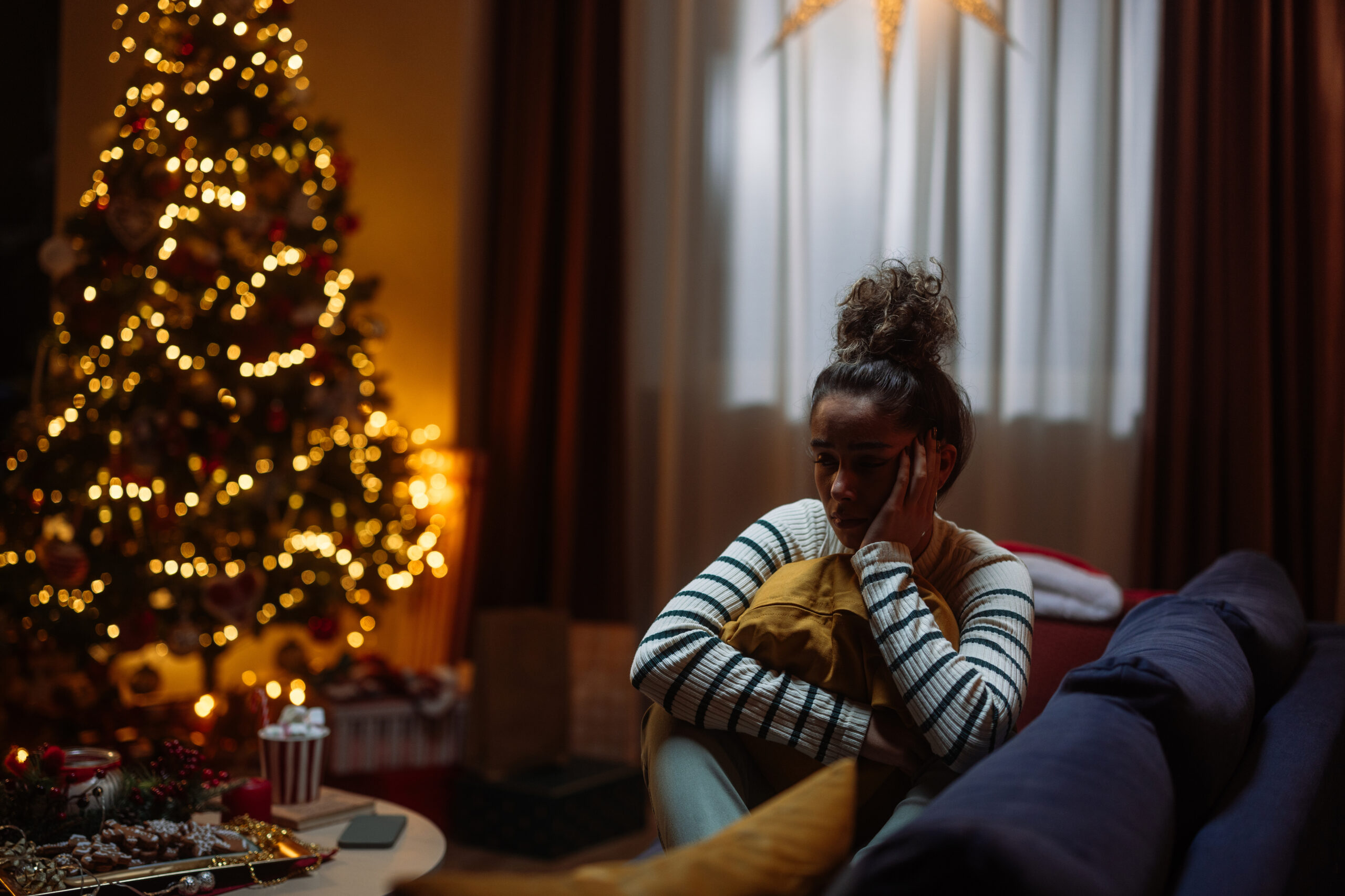 Young woman feeling sad and lonely during christmas time, sitting on the sofa near christmas tree and presents